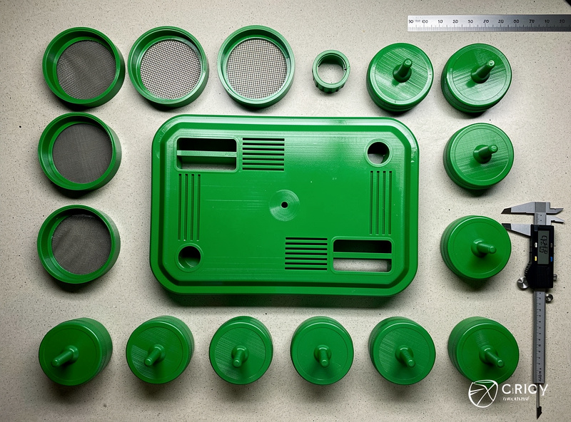 Top-down workshop bench with assorted 3D-printed mushroom cultivation parts laid out monotub lid filter holders jar lids with self healing injection ports all in green PETG