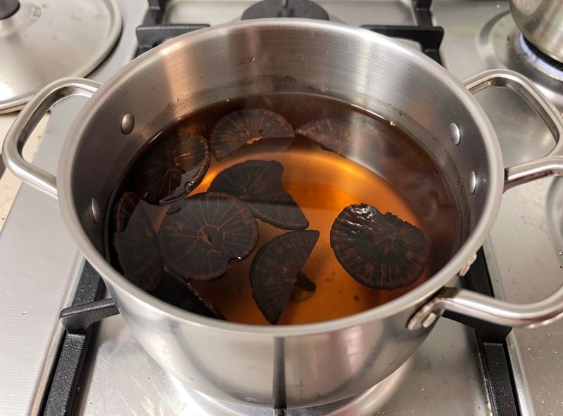 Stainless pot of dark reishi tea at a bare simmer on a stovetop with sliced reishi in the amber liquid